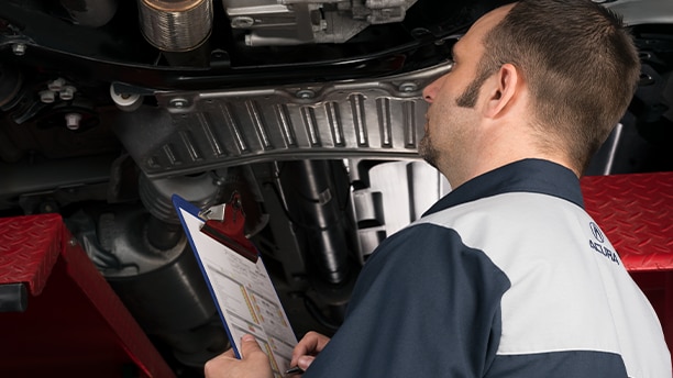 An image showing an Acura technician looking under an Acura with a clipboard.