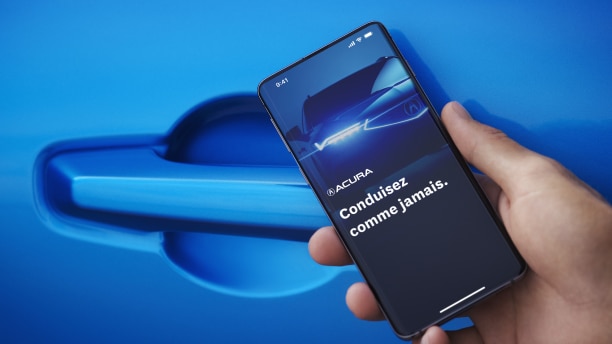 Gros plan d’une main tenant un téléphone intelligent devant la poignée de porte d’une Acura bleue. L’écran du téléphone intelligent affiche une Acura avec les phares allumés et la phrase « Acura. Conduisez comme jamais auparavant. »