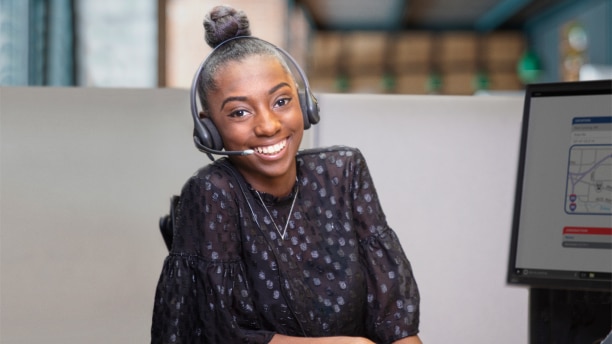 Une jeune femme assise à un poste de travail dans un bureau, avec un casque d’écoute allumé, souriant à la caméra.