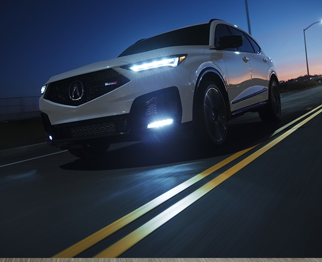A white 2025 MDX Type S drives on an illuminated city highway at night with headlights on.
